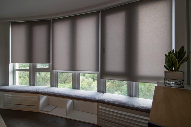 Motorized roller shades in the interior. Automatic roller blinds beige color on big glass windows. Home luxury curtaines are above the windosill with pillows. Electric blinds for home automation.