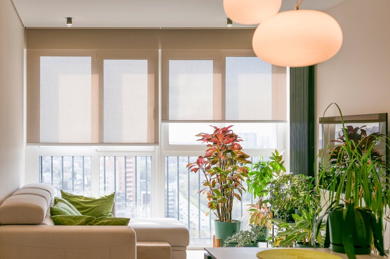 Roller blinds cover large windows in a bright, modern living room filled with lush green plants. Motorized sunscreen shades on the high floor, smart home connected. Screen fabric curtains.