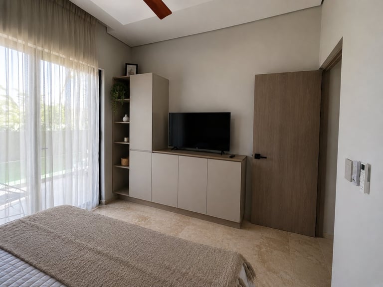Modern bedroom with built-in wooden TV cabinet, white shelving, area rug, and large windows with sheer curtains overlooking outdoor area